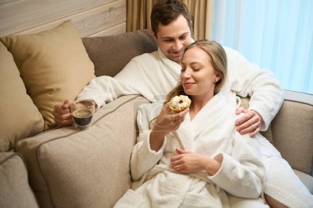 Foto de Hombre sonriente en albornoz bebiendo café y viendo a su novia comiendo sabroso pastel apoyado en él, pareja almorzando sentada en el sofá en casa - Imagen libre de derechos