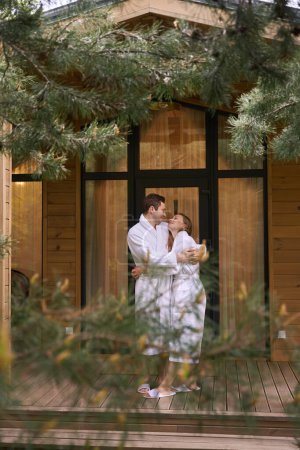 Téléchargez les photos : Mariés couple câlins, respirer l'air frais sur le porche de la maison en bois qu'ils louent en lune de miel, profiter des paysages et les uns des autres, l'amour dans un endroit romantique - en image libre de droit