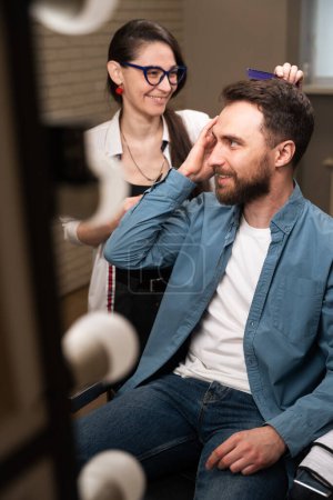 Téléchargez les photos : Barbier femme sur le lieu de travail communique avec un client, un homme en vêtements décontractés assis dans un fauteuil de coiffeur - en image libre de droit