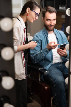 Foto de Mujer joven peluquero en el lugar de trabajo se comunica con un cliente, un hombre muestra el peinado deseado en el teléfono - Imagen libre de derechos