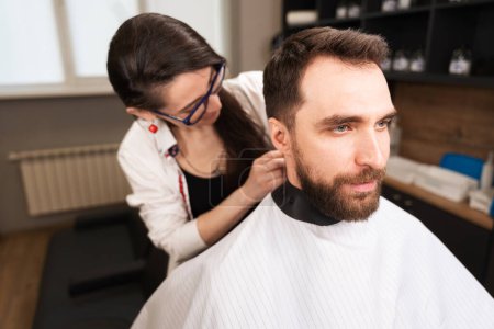 Photo for Woman hairdresser puts on a protective cape to a bearded man, people are reflected in the mirror - Royalty Free Image