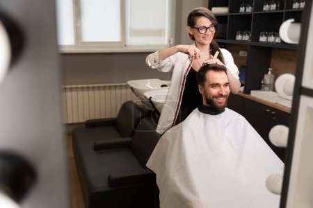 Téléchargez les photos : Jeune femme dans un salon de coiffure coupe les cheveux d'un client avec des ciseaux sous un peigne, les gens se reflètent dans le miroir - en image libre de droit