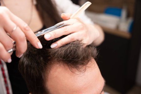 Foto de Hombre adulto se sienta en una silla de peluquero, el maestro corta el pelo de los clientes con tijeras debajo del peine - Imagen libre de derechos