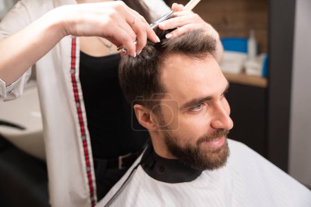 Photo for Smiling man sits in a barber chair, the master cuts the client with scissors under the comb - Royalty Free Image