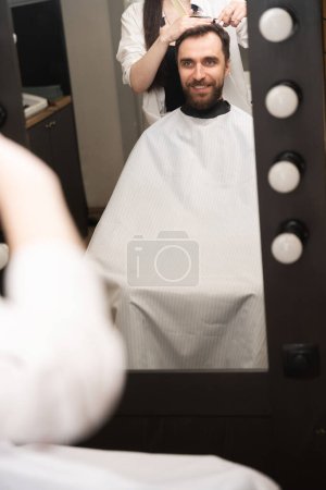 Foto de Maestro en la peluquería corta el pelo de los clientes con tijeras debajo del peine, la gente se refleja en el espejo - Imagen libre de derechos