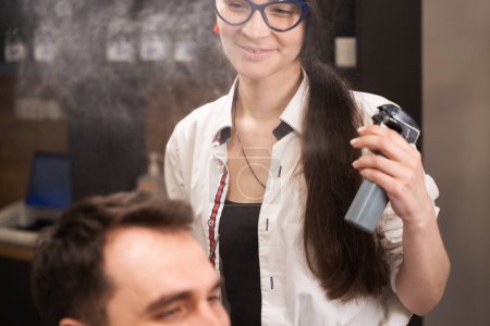 Téléchargez les photos : Jolie femme dans des lunettes et une boucle d'oreille rouge coiffe les cheveux des clients, elle utilise un spray coiffant spécial - en image libre de droit