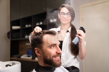 Téléchargez les photos : Barbier femme dans un salon de coiffure coiffe un cheveux clients, elle utilise un spray coiffant spécial - en image libre de droit