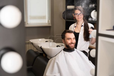 Téléchargez les photos : Mignon coiffeur femme dans coiffeur cheveux masculins coiffure, elle utilise spray coiffure - en image libre de droit