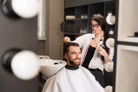 Téléchargez les photos : Jeune femme coiffeur coiffant les cheveux des hommes, elle utilise un spray coiffant et peigne - en image libre de droit