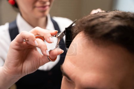 Téléchargez les photos : Maître coiffeur coupe un client sous un peigne avec des ciseaux, une femme en uniforme - en image libre de droit