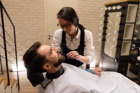 Téléchargez les photos : Femme barbier à lunettes coupe la barbe des clients, l'homme s'assoit dans une chaise confortable - en image libre de droit