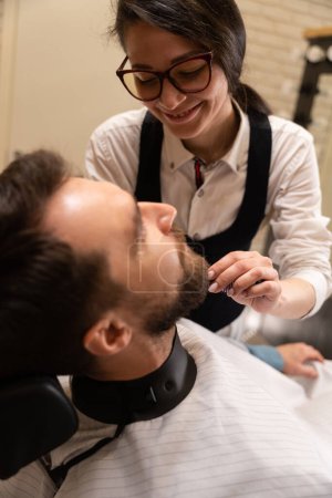 Téléchargez les photos : Jeune femme barbier coupe une barbe à un client, l'homme s'assoit dans une chaise confortable - en image libre de droit