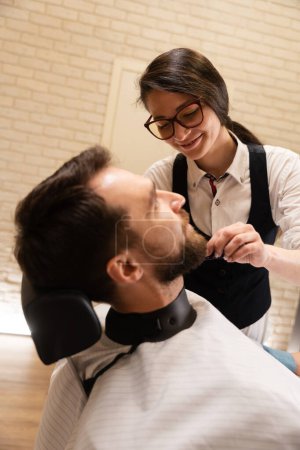 Foto de Linda hembra peluquero corta una barba a un cliente, el hombre se encuentra en una silla cómoda - Imagen libre de derechos