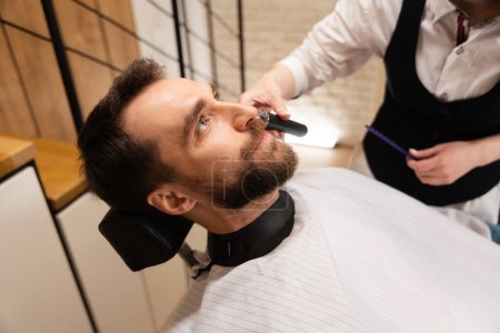 Photo for Barber cuts the beard and mustache to the client, the man is sitting in a comfortable chair - Royalty Free Image