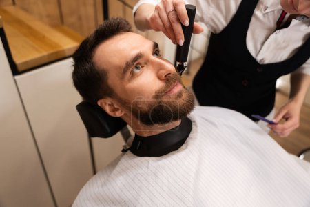Foto de Peluquero corta la barba y el bigote al cliente, el maestro utiliza un cortador de barba especial - Imagen libre de derechos