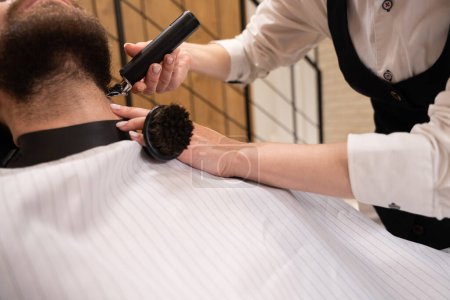 Foto de Peluquero femenino en el lugar de trabajo corta la barba de un hombre, el maestro utiliza un cortador de barba especial - Imagen libre de derechos