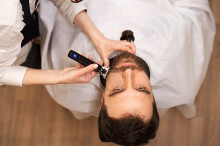Téléchargez les photos : Maître dans le salon de coiffure coupe la barbe d'un homme, la femme utilise une tondeuse à barbe spéciale - en image libre de droit
