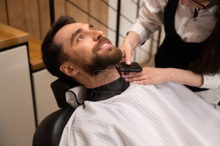 Téléchargez les photos : Homme client dans un salon de coiffure sur une coupe de barbe, le maître utilise des outils spéciaux - en image libre de droit