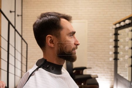 Photo for Bearded man sits in front of a mirror in a barbershop, a client in a protective cape - Royalty Free Image
