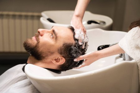 Foto de Hombre guapo disfruta de procedimiento de lavado de cabello en la peluquería, peluquería utiliza champú especial - Imagen libre de derechos