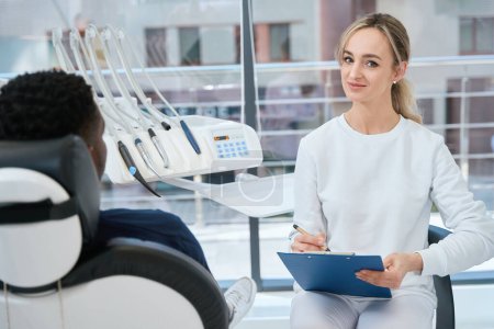 Photo for Young woman dental technician holding medical records, making notes listening to male client, posing, looking at camera, private dental care clinic - Royalty Free Image