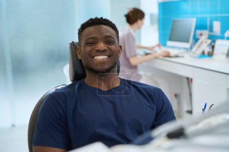 Téléchargez les photos : Client masculin assis dans une chaise dentaire avec un sourire sain et propre tout en stomatologue tenant des dossiers médicaux sur ordinateur - en image libre de droit