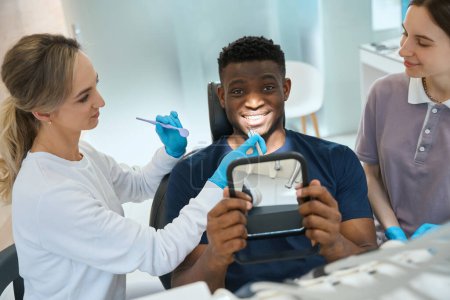 Photo for Qualified female dentist and nurse working on teeth health of African American client sitting in dental chair and holding mirror to see healthy teeth - Royalty Free Image