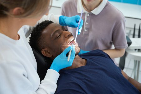Téléchargez les photos : Stomatologue femelle regardant miroir buccal vérifier la santé des dents des jeunes hommes, check-up et traitement de routine, nettoyage des dents professionnel - en image libre de droit