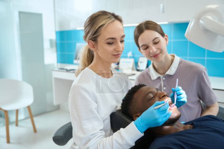Téléchargez les photos : Jeune infirmière assistant la femme dentiste lors du bilan de santé de l'homme afro-américain cavité buccale, prévention des problèmes dentaires, soins de santé - en image libre de droit