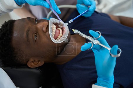 Foto de Cliente afroamericano ampliamente abierto boca y mujer que espera médico haciendo inyección anestésica, cita con el estomatólogo, primer plano - Imagen libre de derechos