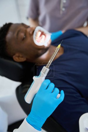 Foto de Paciente asustado con retractor bucal sentado en la silla dental y mirando el inyector de anestesia local eléctrica, estomatólogo preparándose para la inyección de anestesia - Imagen libre de derechos