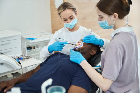 Foto de Enfermera cuidadora sosteniendo eyector de saliva mientras la mujer dentista usa inyector de anestesia oral, cuidado estomatológico para paciente afroamericano con dolor de muelas - Imagen libre de derechos