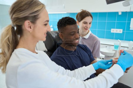 Foto de Mujer dentista mostrando a afroamericano el trabajo del cliente masculino y la vista del puente dental, enfermera calificada y médico explicando la forma de prótesis - Imagen libre de derechos