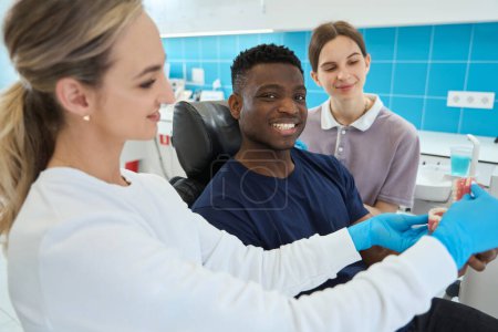 Téléchargez les photos : Femme technicienne dentaire montrant à quoi ressemblent les fausses dents ou les facettes sur le modèle de mâchoire 3D, client afro-américain intéressant dans les services de dentisterie esthétique - en image libre de droit