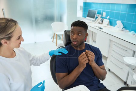 Photo for Smiling woman dentist calming down scared young African American man who coming to doctor appointment and sitting in dental chair - Royalty Free Image