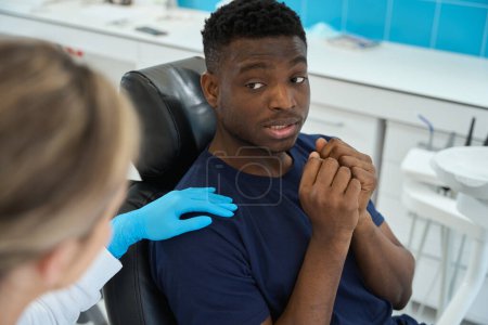 Foto de Paciente varón asustado sentado en silla de ortodoncia y pidiendo al médico femenino que no lo lastime, miedo al pánico de los dentistas, médico amigable calmando al hombre - Imagen libre de derechos