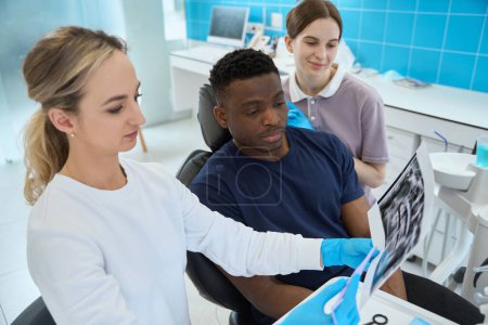 Téléchargez les photos : Femme stomatologue focalisée regardant l'image radiographique du client masculin, à la recherche de dents et d'abcès problématiques, infirmière va aider pendant le traitement - en image libre de droit