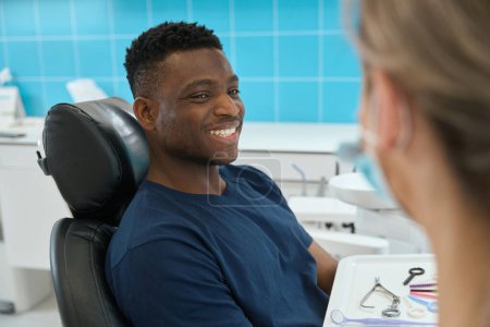 Photo for Cheerful African American man sitting in dental chair and looking at high-qualified woman stomatologist with toothy smile, satisfied with private clinic service - Royalty Free Image