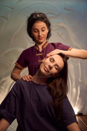 Photo for Spa client sitting with eyes closed while masseuse tilting her head to side - Royalty Free Image
