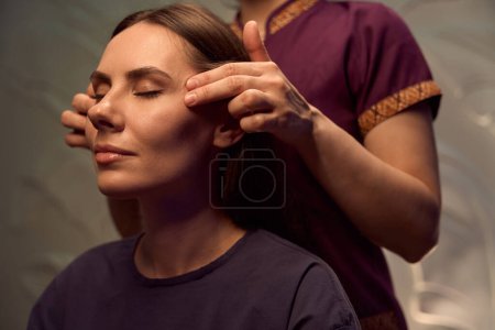 Photo for Relaxed lady sitting with eyes closed while masseuse applying gentle pressure with fingers to her temples - Royalty Free Image