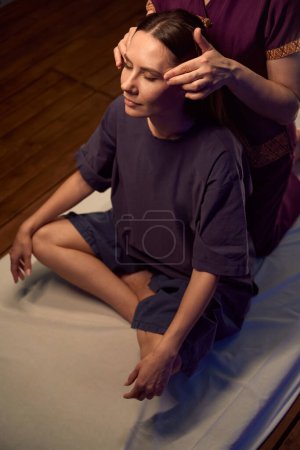 Foto de Mujer sentada con los ojos cerrados y las piernas cruzadas sobre futón mientras masoterapeuta masajea sus sienes en movimientos circulares - Imagen libre de derechos