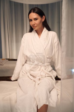 Photo for Relaxed young woman in bathrobe sitting on couch in wellness center and looking before her - Royalty Free Image