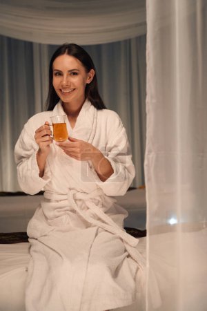 Téléchargez les photos : Jeune femme souriante en peignoir éponge assise sur un canapé de massage avec une tasse de thé dans les mains - en image libre de droit