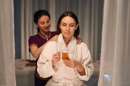 Photo for Young woman sitting with cup of herbal drink during Thai shoulder massage performed by experienced masseuse - Royalty Free Image