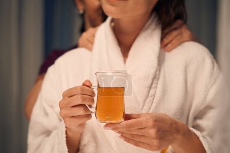 Foto de Foto recortada del cliente del spa sosteniendo una taza de bebida mientras masoterapeuta masajea sus hombros - Imagen libre de derechos