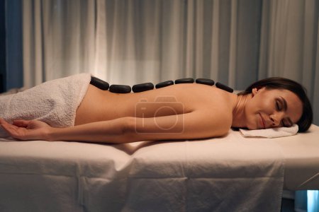 Foto de Vista lateral de la mujer acostada propensa con piedras lisas en su espalda desnuda durante la sesión de masoterapia - Imagen libre de derechos