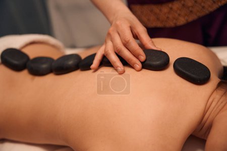 Téléchargez les photos : Photo recadrée du thérapeute spa plaçant des pierres chaudes sur le dos nu du patient - en image libre de droit