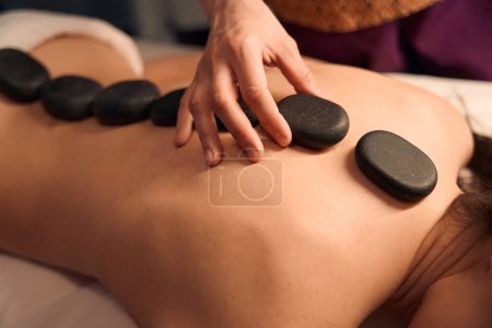 Photo for Cropped photo of masseuse putting heated flat basalt stones along client spine - Royalty Free Image
