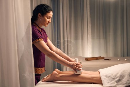 Photo for Side view of focused massotherapist massaging woman legs with pair of herbal balls - Royalty Free Image
