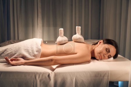 Photo for Young female lying with eyes closed on massage table with herbal pouches on naked back - Royalty Free Image
